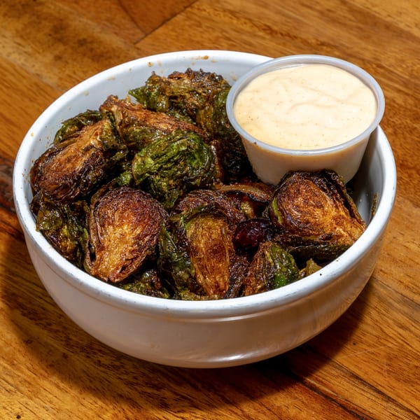 A white bowl filled with crispy, dark green roasted Brussels sprouts and a side of creamy dipping sauce.